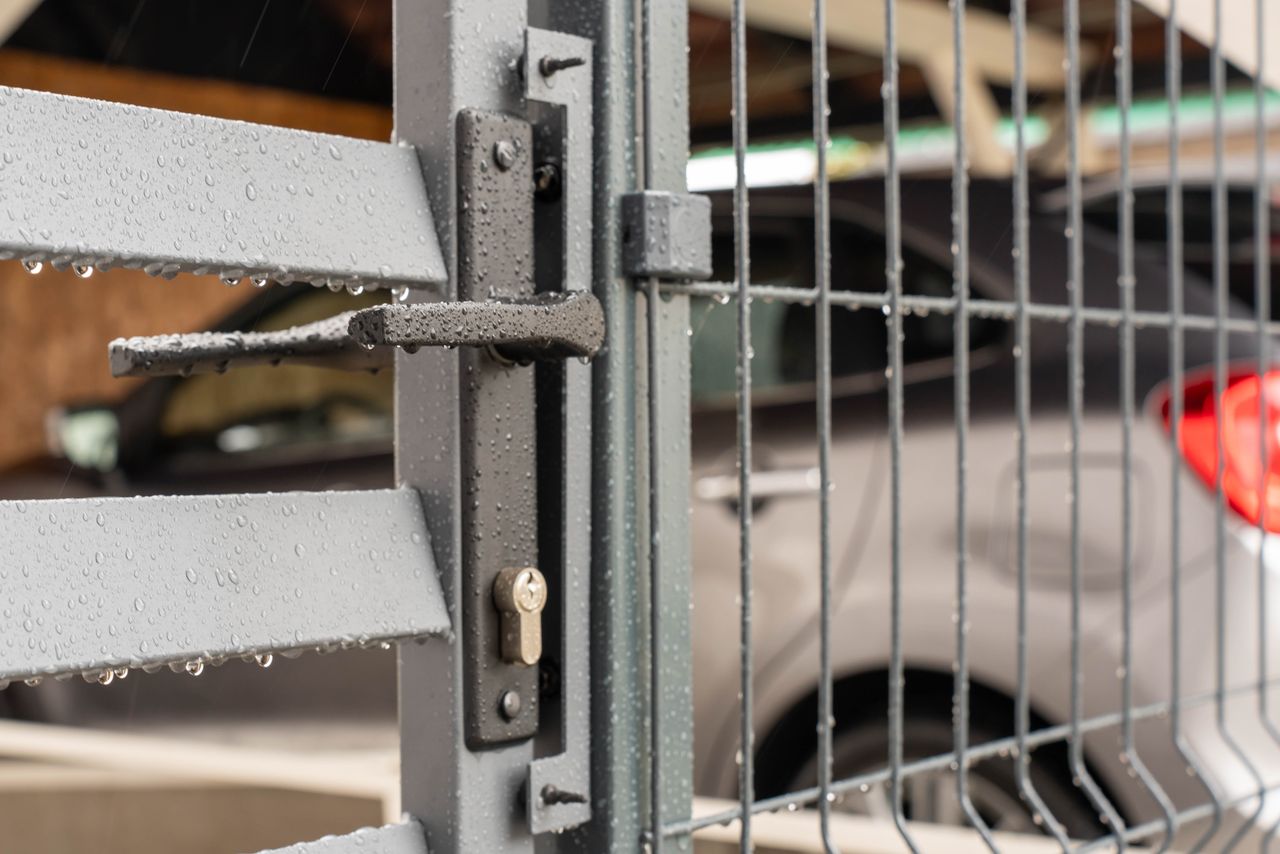 Detailed view of a wet metal gate with raindrops and a parked car in the background.